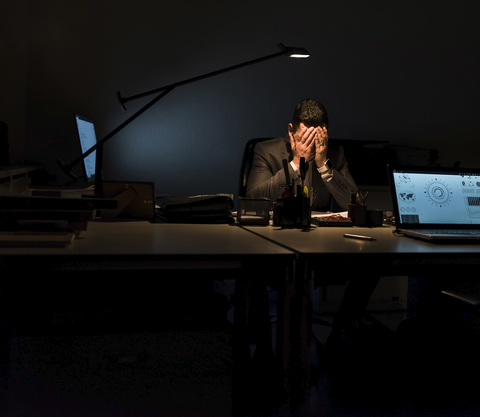 Überforderter Geschäftsmann sitzt im Dunkeln an seinem Schreibtisch, lizenzfreies Stockfoto