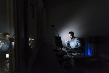 Geschäftsmann mit einer Tasse Kaffee bei der Arbeit am Computer im Büro bei Nacht - UUF13216