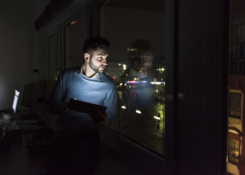 Businessman sitting with tablet on window sill in office at night looking out of the window - UUF13229