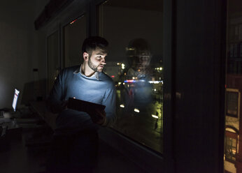 Geschäftsmann sitzt mit Tablet auf der Fensterbank im Büro und schaut nachts aus dem Fenster - UUF13229