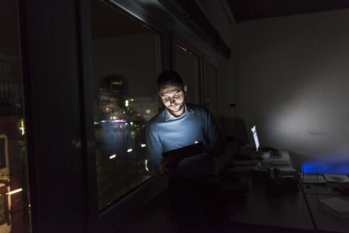 Geschäftsmann sitzt nachts auf der Fensterbank im Büro und benutzt ein Tablet - UUF13230
