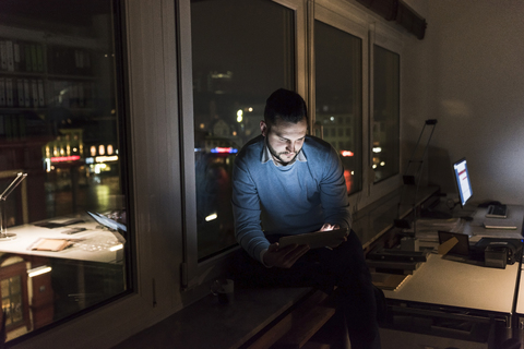 Geschäftsmann sitzt nachts auf der Fensterbank im Büro und benutzt ein Tablet, lizenzfreies Stockfoto