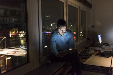 Businessman sitting on window sill in office at night using tablet - UUF13231