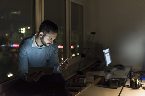 Businessman sitting on windowsill in office at night looking at smartphone - UUF13232