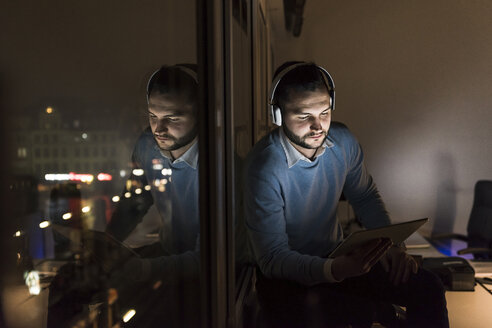 Geschäftsmann sitzt nachts auf der Fensterbank im Büro und benutzt Tablet und Kopfhörer - UUF13233