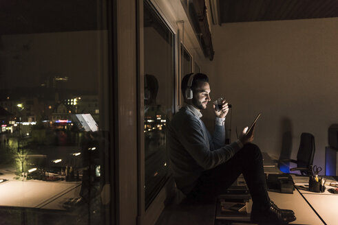 Geschäftsmann sitzt nachts auf der Fensterbank im Büro und hört Musik mit Kopfhörern - UUF13235