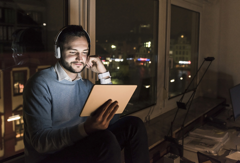 Businessman sitting on window sill in office at night using tablet and headphones stock photo