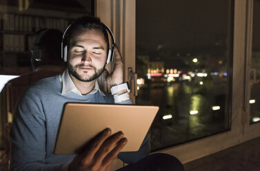 Businessman sitting on window sill in office at night listening music with headphones and tablet - UUF13237