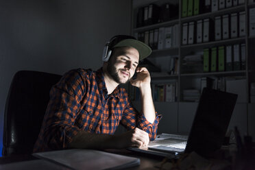 Porträt eines Freiberuflers, der nachts am Schreibtisch sitzt und Laptop und Kopfhörer benutzt - UUF13245