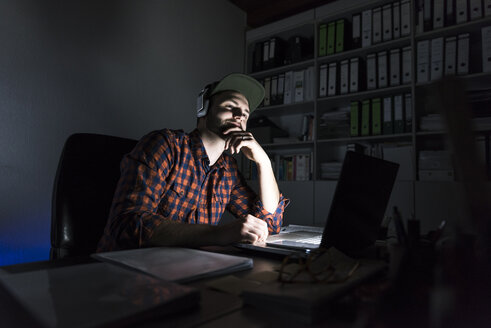 Porträt eines Freiberuflers, der nachts am Schreibtisch sitzt und Laptop und Kopfhörer benutzt - UUF13246
