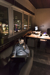 Geschäftsmann im Büro, der nachts mit Laptop und Kopfhörern auf der Couch sitzt - UUF13254