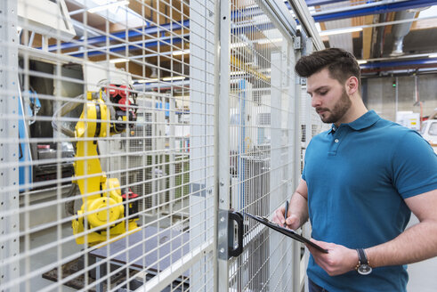 Mann mit Klemmbrett steht vor Industrieroboter in moderner Fabrik - DIGF03833