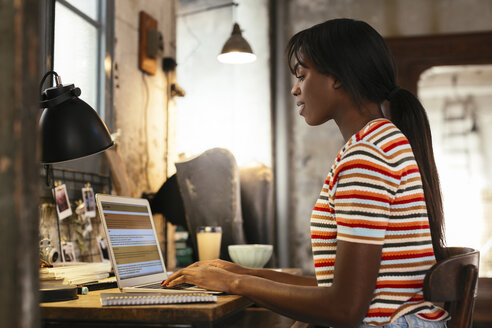 Junge Frau sitzt am Schreibtisch in einem Loft und arbeitet am Laptop - EBSF02281