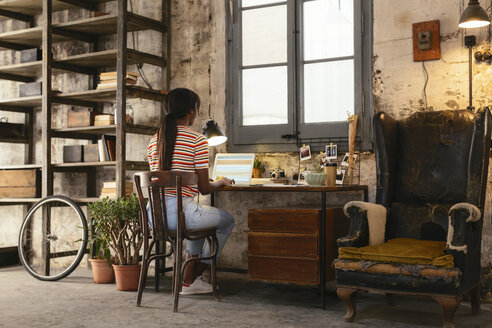 Rückenansicht einer jungen Frau, die an einem Schreibtisch in einem Loft sitzt und an einem Laptop arbeitet - EBSF02283