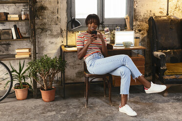 Young woman sitting in front of a desk in a loft using cell phone - EBSF02285