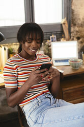 Porträt einer lächelnden jungen Frau, die in einem Loft vor einem Schreibtisch sitzt und ein Mobiltelefon benutzt - EBSF02286