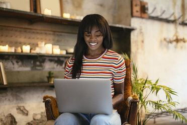 Porträt einer lächelnden jungen Frau, die auf einem Lederstuhl sitzt und einen Laptop benutzt - EBSF02299