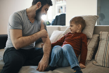 Vater auf der Couch sitzend mit kleinem Sohn, der sein Tattoo zeigt - ZEDF01288