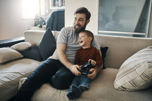 Happy father and son sitting together on the couch playing computer game - ZEDF01302