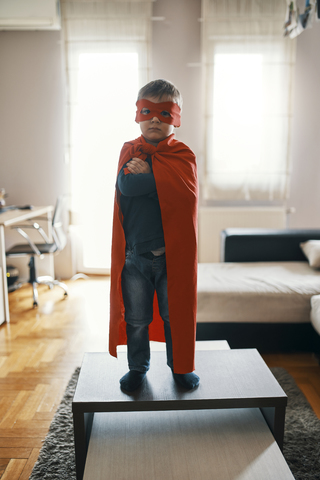 Kleiner Junge als Superheld verkleidet auf dem Kaffeetisch zu Hause stehend, lizenzfreies Stockfoto