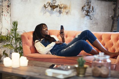 Lachende Frau, die zu Hause auf der Couch liegt und ein Mobiltelefon benutzt, lizenzfreies Stockfoto