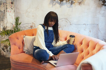 Young woman sitting on the couch with coffee to go in a loft using laptop - EBSF02325