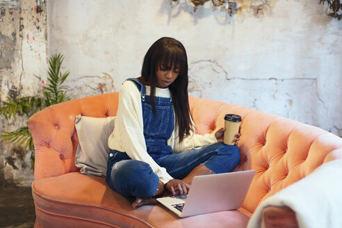 Junge Frau sitzt auf der Couch mit Kaffee zum Mitnehmen in einem Loft und benutzt einen Laptop - EBSF02325
