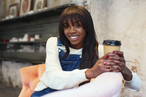 Porträt einer lächelnden Frau mit Kaffee zum Mitnehmen, die auf der Couch in einem Loft sitzt - EBSF02326