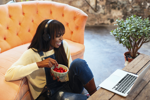 Lachende junge Frau, die mit Laptop und Kopfhörern auf dem Boden ihres Lofts sitzt, lizenzfreies Stockfoto