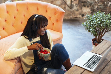 Laughing young woman sitting on the floor of her loft using laptop and headphones - EBSF02333