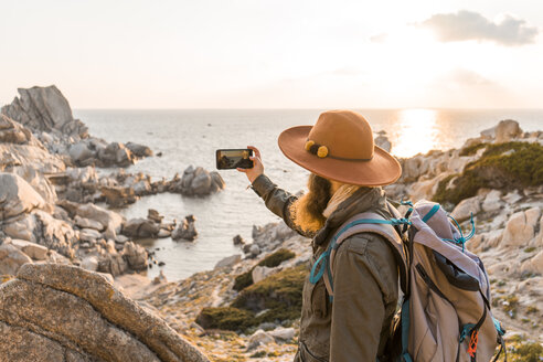 Italien, Sardinien, Wanderer mit Hut und Rucksack fotografiert das Meer mit dem Mobiltelefon - AFVF00410