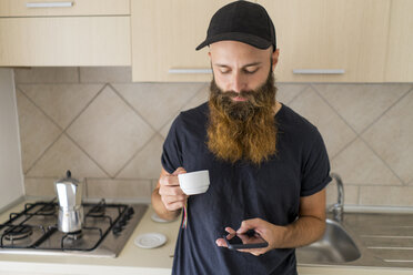 Bärtiger Mann steht mit Espressotasse in der Küche und schaut auf sein Handy - AFVF00429