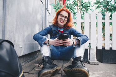 Porträt einer rothaarigen jungen Frau, die ein Mobiltelefon benutzt, während sie an der Tür sitzt - MASF01366