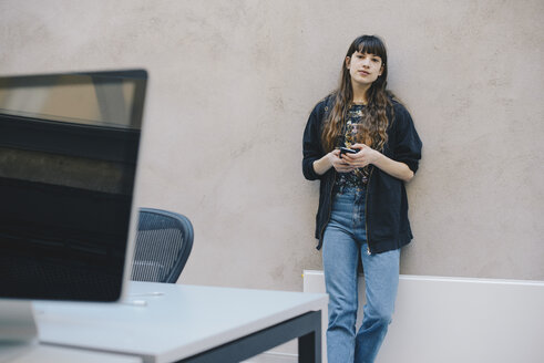 Porträt eines weiblichen Computerprogrammierers, der ein Smartphone hält und an einer beigen Wand im Büro steht - MASF01375