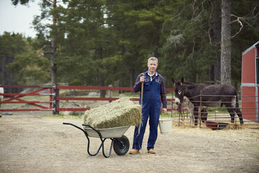 Portrait of farmer holding bucket and pitchfork while standing by wheelbarrow in farm - MASF01385