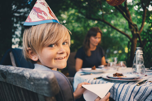 Porträt eines glücklichen Jungen mit Hut und Seidenpapier in der Hand bei einem Abendessen im Garten - MASF01390