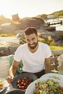 Porträt eines glücklichen jungen Mannes, der das Mittagessen auf einer Party auf einem Steg genießt, während eines sonnigen Tages - MASF01401