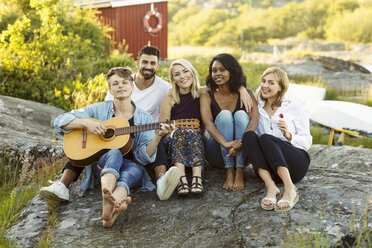 Portrait of man holding guitar sitting with friends on rock formation - MASF01402