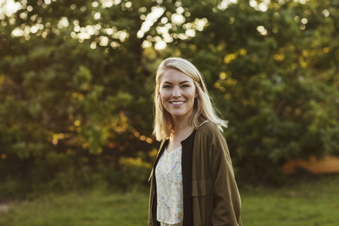 Porträt einer lächelnden jungen blonden Frau, die vor einem Baum steht, lizenzfreies Stockfoto