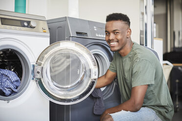 Porträt eines lächelnden jungen Mannes, der vor einer Waschmaschine im Waschsalon kniet - MASF01423