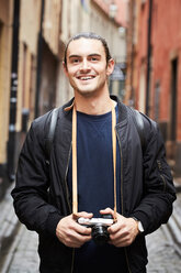 Porträt eines lächelnden jungen Mannes, der eine Kamera hält und in einer Gasse steht - MASF01447