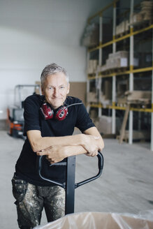 Portrait of mature worker leaning on pallet jack by stack at industry - MASF01458