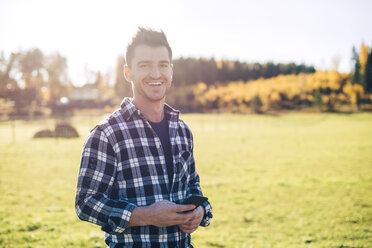 Portrait of smiling mid adult male farmer holding mobile phone while standing on field - MASF01463