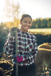 Porträt einer lächelnden erwachsenen Bäuerin, die eine Schaufel hält, mit Schweinen im Hintergrund auf einem Bio-Bauernhof - MASF01464