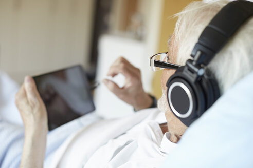 Blick über die Schulter eines älteren Mannes, der Kopfhörer trägt, während er ein digitales Tablet im Krankenhaus benutzt - MASF01477