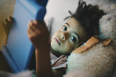 Porträt eines Jungen, der ein digitales Tablet benutzt, während er mit einem Teddybären zu Hause auf dem Bett liegt - MASF01481