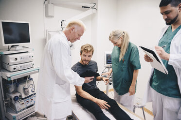 Reifer Arzt, der einem jungen männlichen Patienten auf dem Bett von Kollegen im Krankenhaus ein digitales Tablet zeigt - MASF01552