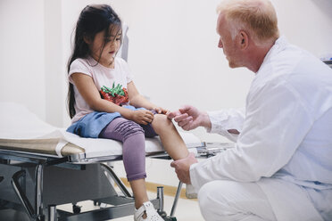 Mature doctor examining girl's knee sitting on bed at hospital - MASF01555