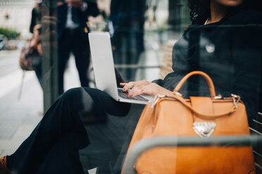 Mittelteil einer Geschäftsfrau, die einen Laptop benutzt und in einem Wartehäuschen sitzt, gesehen von einem Glas - MASF01592
