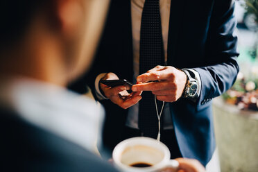 Mittelteil eines Geschäftsmannes, der sein Smartphone benutzt, während er mit einem Kollegen in einem Café Kaffee trinkt - MASF01599